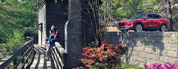 two people hang out on a bridge in a grassy area with a red ford edge parked on a hill behind them 