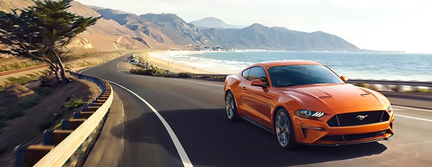 an orange ford mustang drives down a road next to a beach with mountains in the background