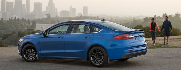 a blue ford fusion sits on a road overlooking a cityscape while a couple walks towards it hand in hand