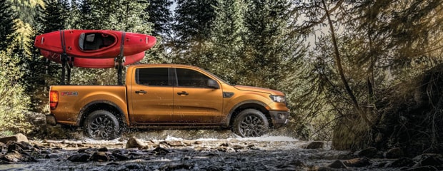 an orange ford ranger that has a red canoe strapped to the bed drives through a small stream in a forest