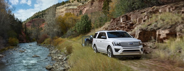 a white ford expedition drives in a mountain area alongside a stream. it is towing a blue ATV