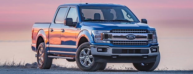 a blue ford truck sits on a grassy area at sunset with a pink sky in the background 