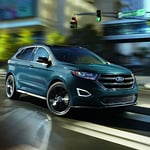 a blue ford edge sits in the middle of an intersection under a light with a blurry dark background
