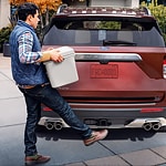 a man carrying a cooler extends his foot towards the trunk of a red ford explorer 